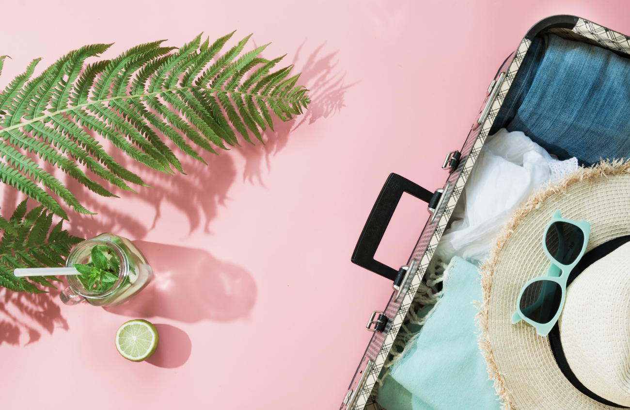 pink backdrop calling you to travel, an open suitcase with clothes, sunglasses, sunhat, and palm branches paid out with a fun cocktail.