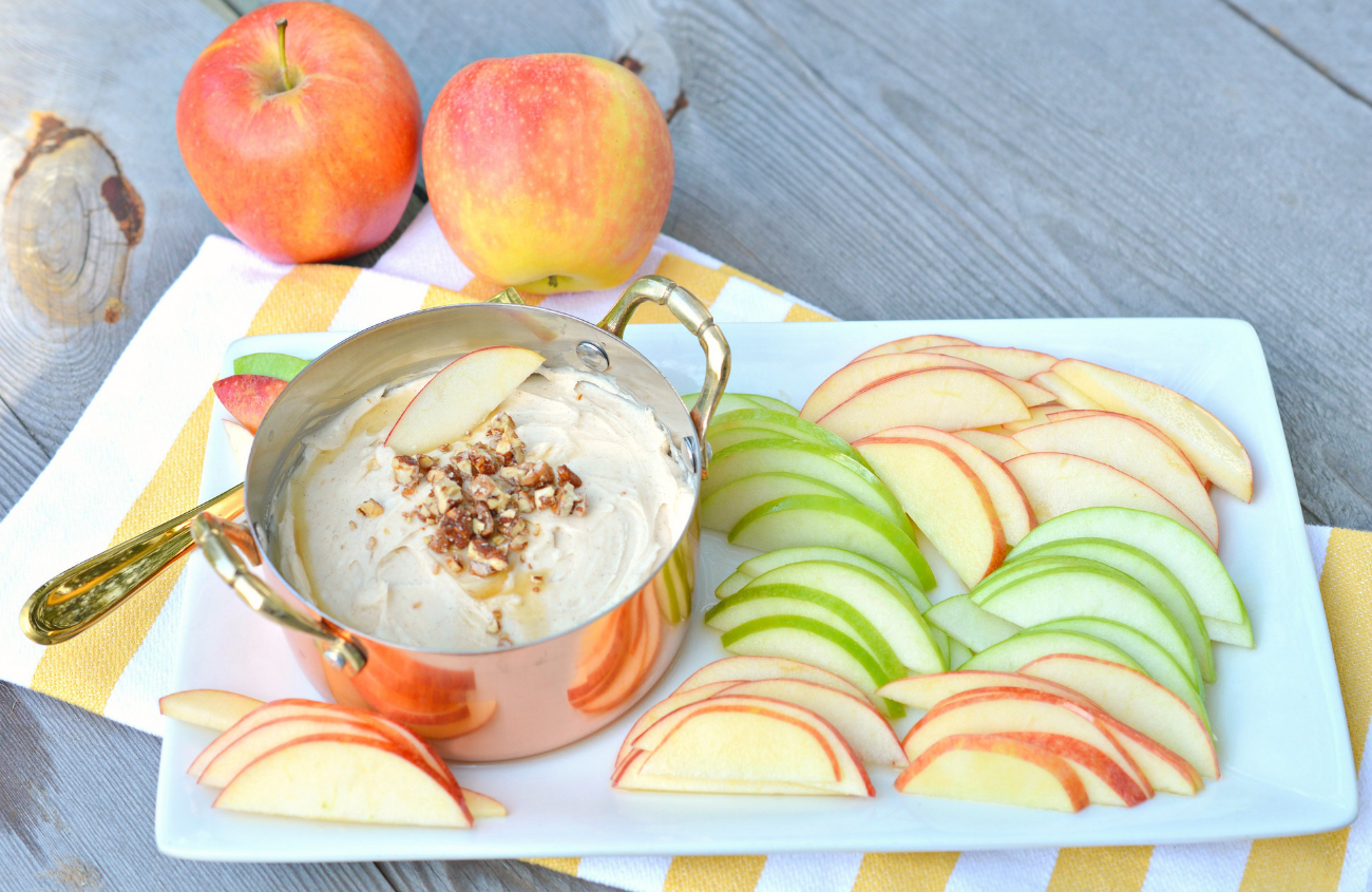 Horizontal image of a pan filled with caramel apple cream cheese dip on a white tray and surrounded by green and red apple slices.